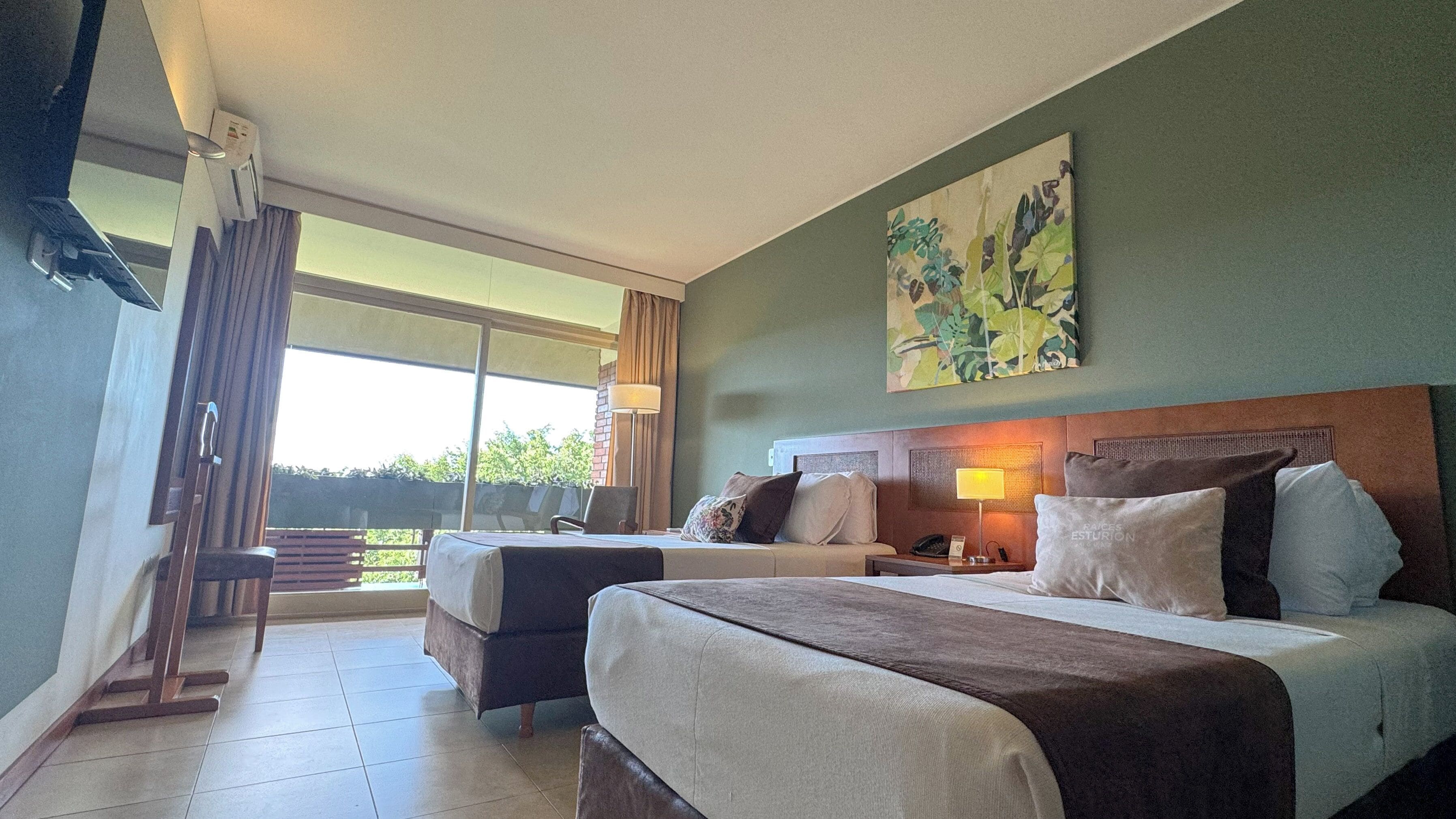 Habitación de hotel con dos camas, un ventanal grande, un cuadro en la pared y vistas al exterior con vegetación.