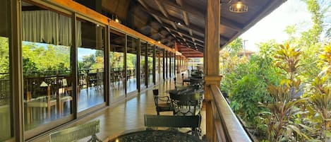 An outdoor covered patio with tables and chairs, large windows, and a view of greenery.