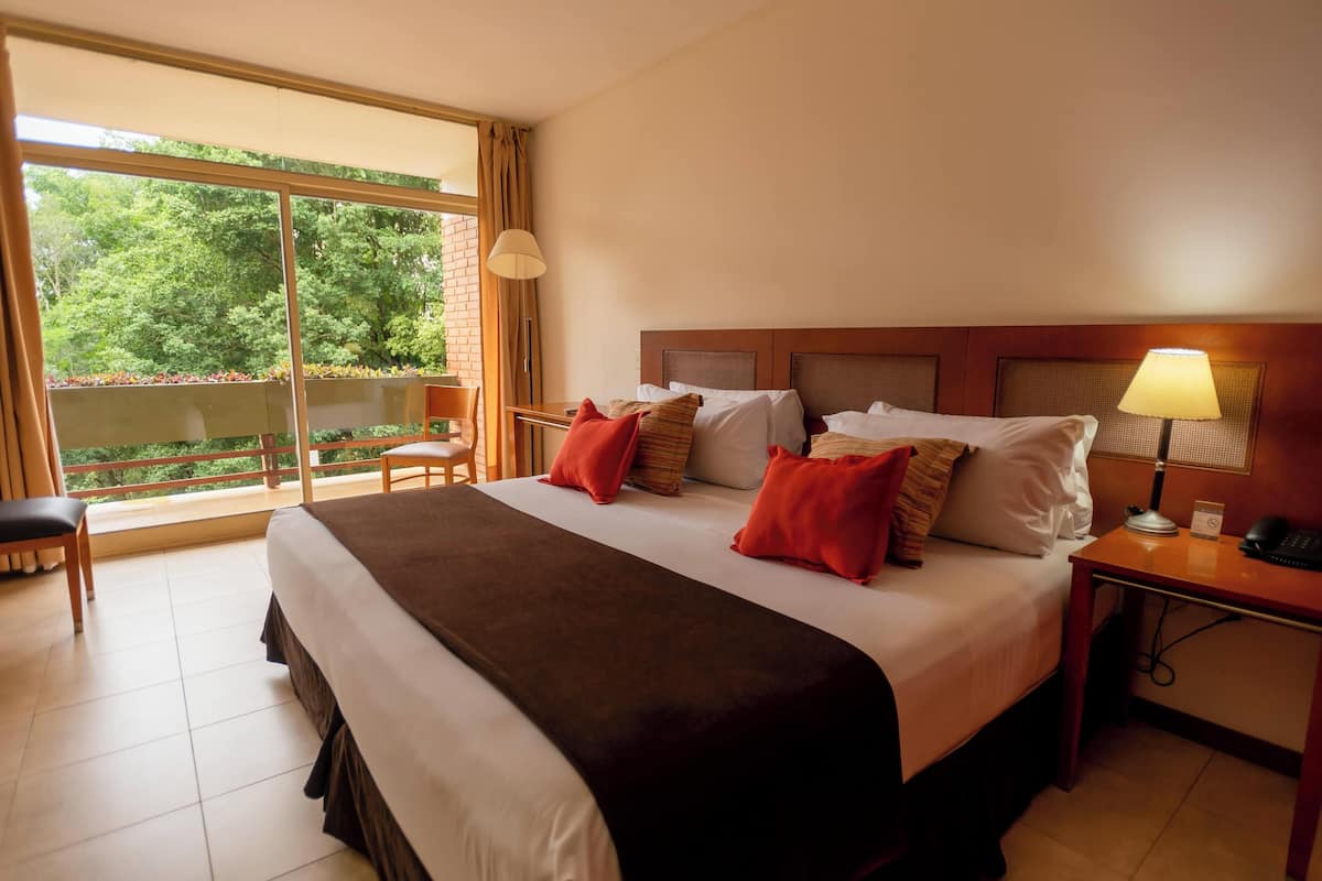 Une chambre d’hôtel avec un grand lit, un bureau, une chaise et un balcon donnant sur la verdure.