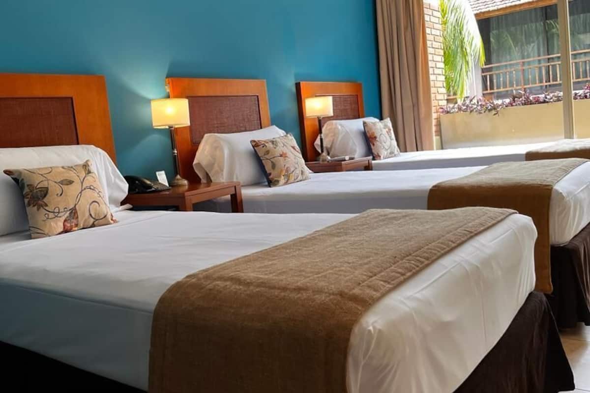 Une chambre d’hôtel avec trois lits, chacun doté d’une tête de lit en bois, de draps blancs et d’oreillers décoratifs. On y trouve des tables de chevet avec des lampes, ainsi qu’une fenêtre donnant sur un paysage verdoyant.