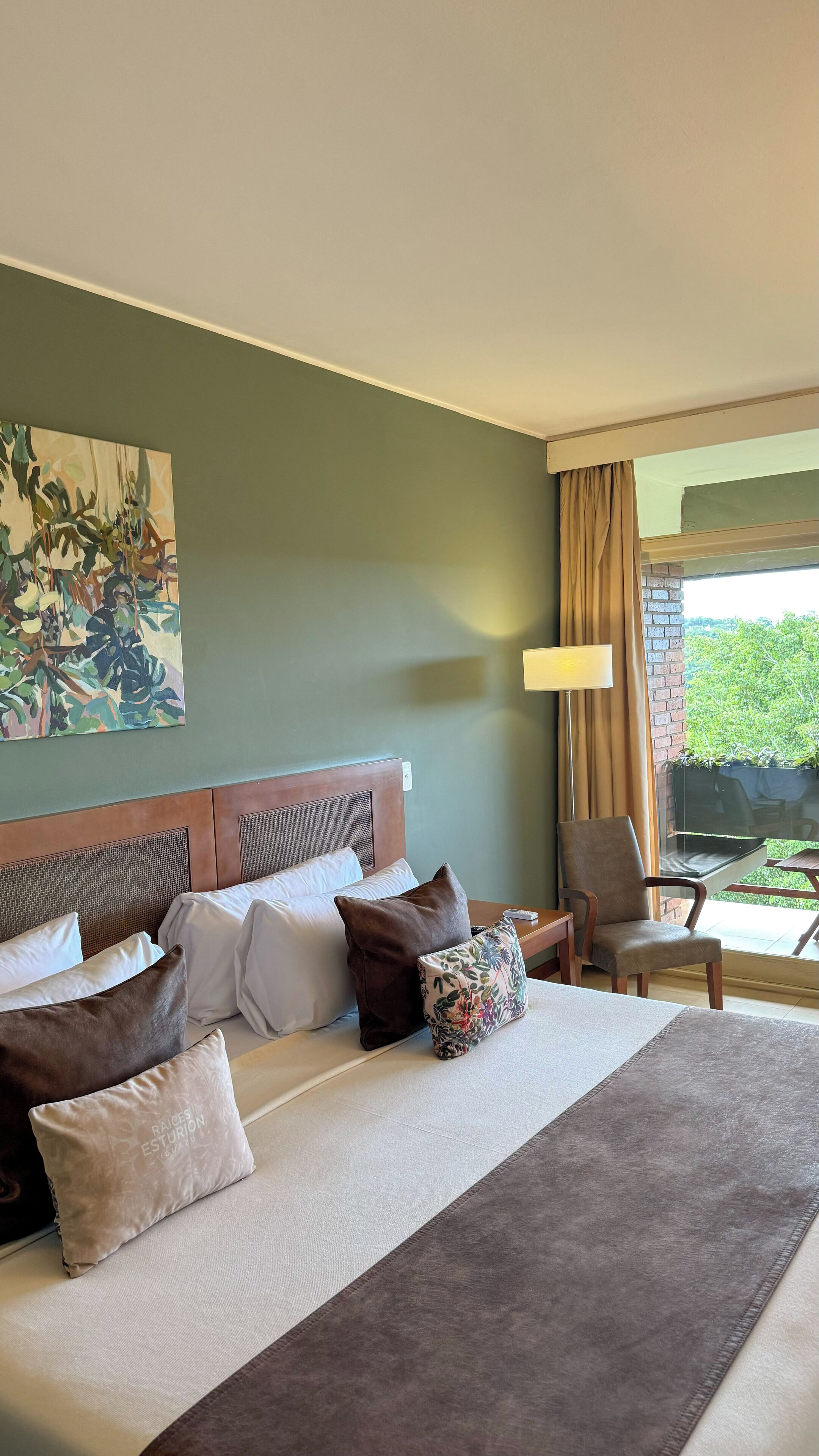Ett hotellrum med en stor säng, flera kuddar, ett konstverk på väggen, en stol och utsikt över grönområden utanför.