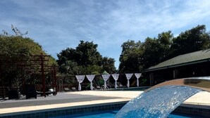 Una piscina con fuente, rodeada de árboles y una zona cubierta con sombrillas.