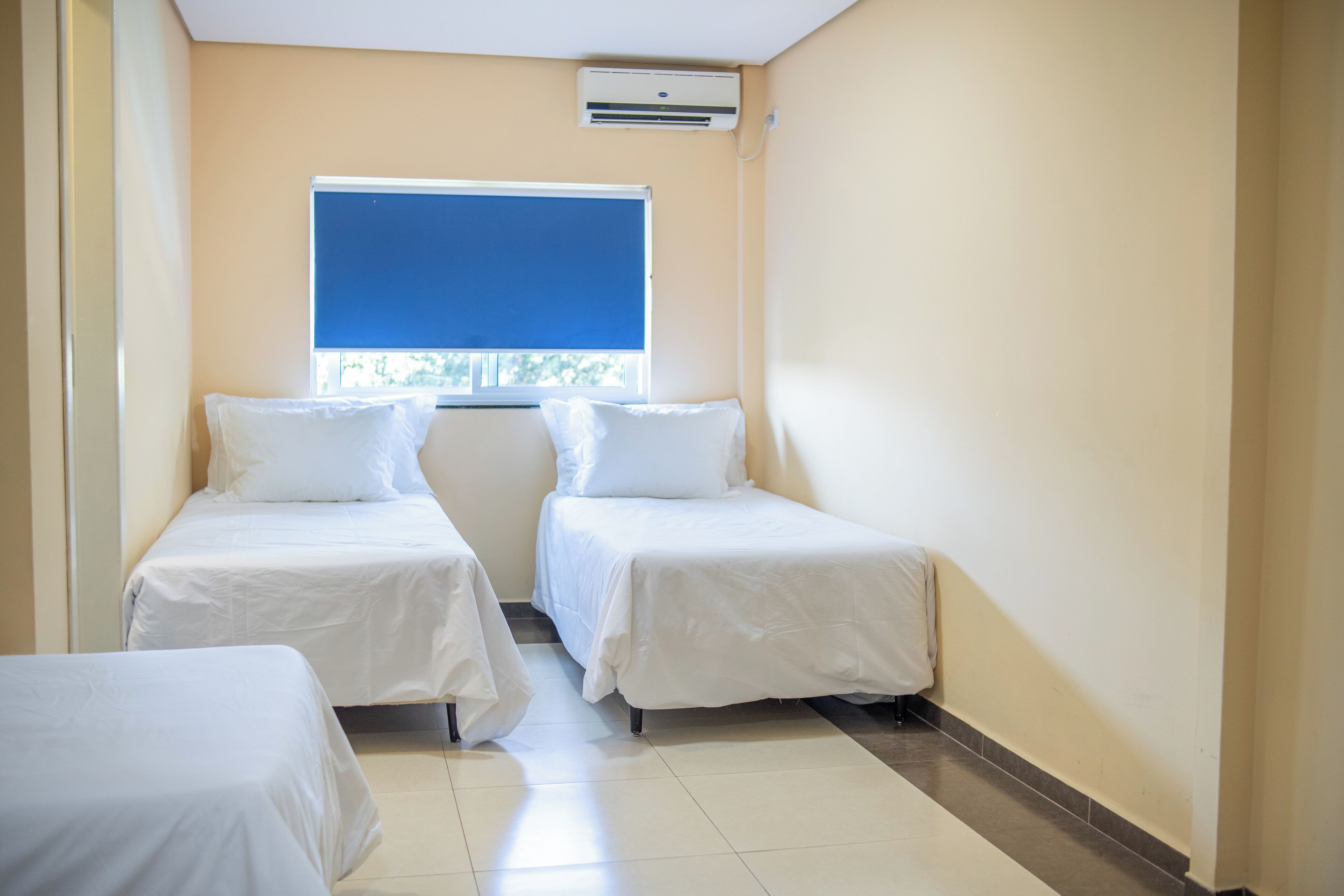 Ein Zimmer mit zwei Einzelbetten, einem Fenster mit blauer Jalousie und einer Klimaanlage an der Wand.