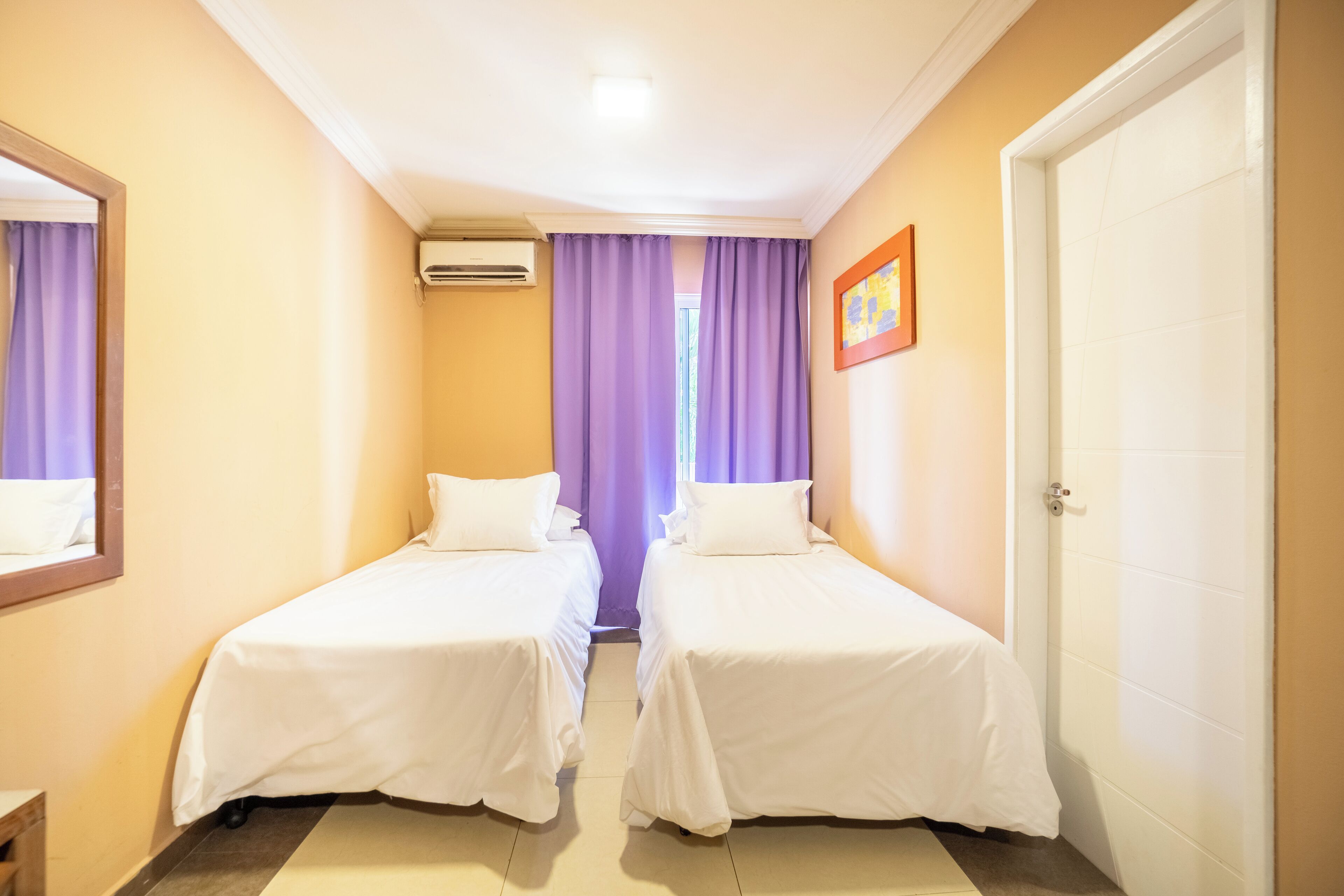 Ett hotellrum med två separata sängar, en dörr, en luftkonditioneringsanläggning och ett fönster med lila gardiner.