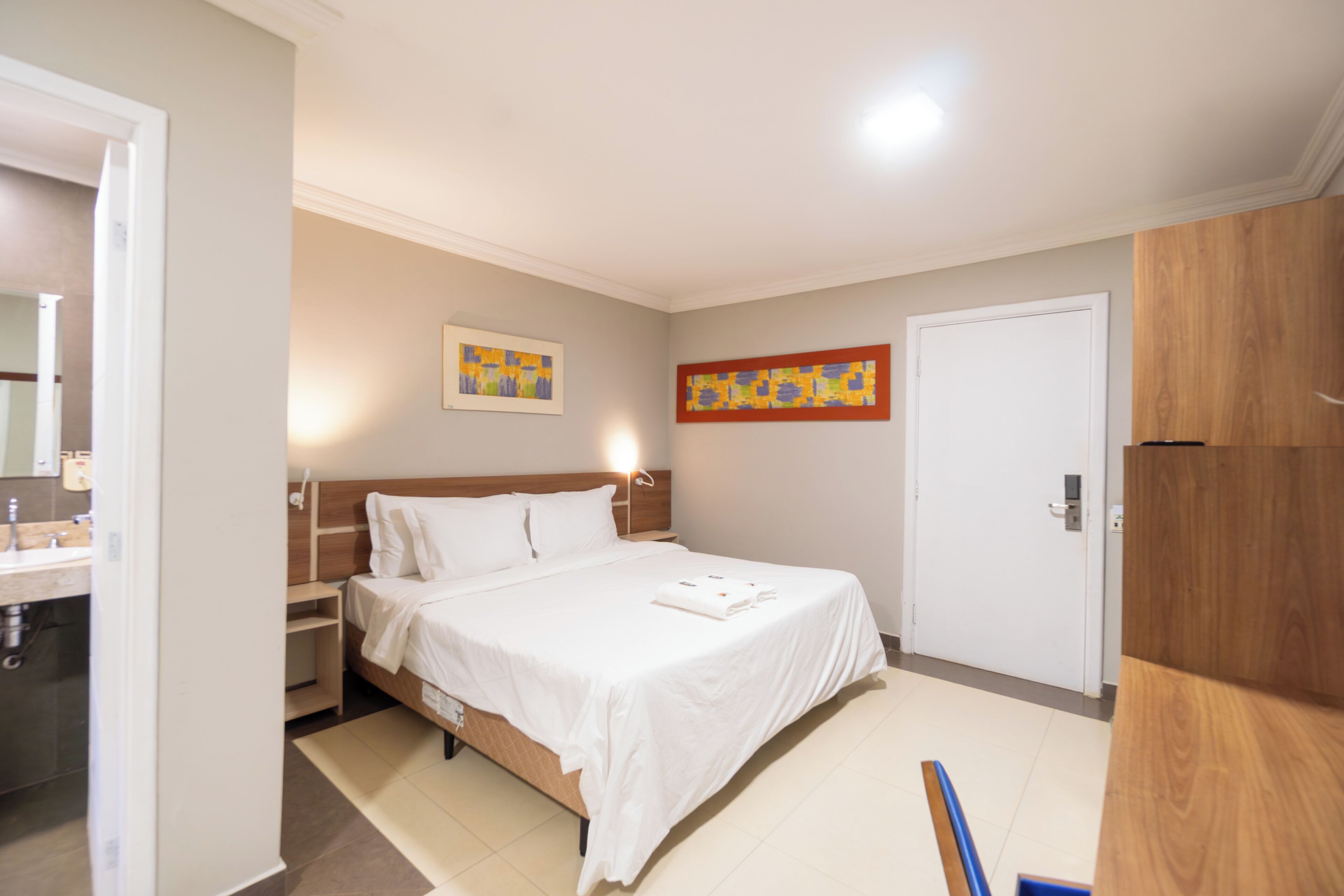 Habitación de hotel con cama, armario de madera, baño con lavamanos y dos cuadros en la pared.