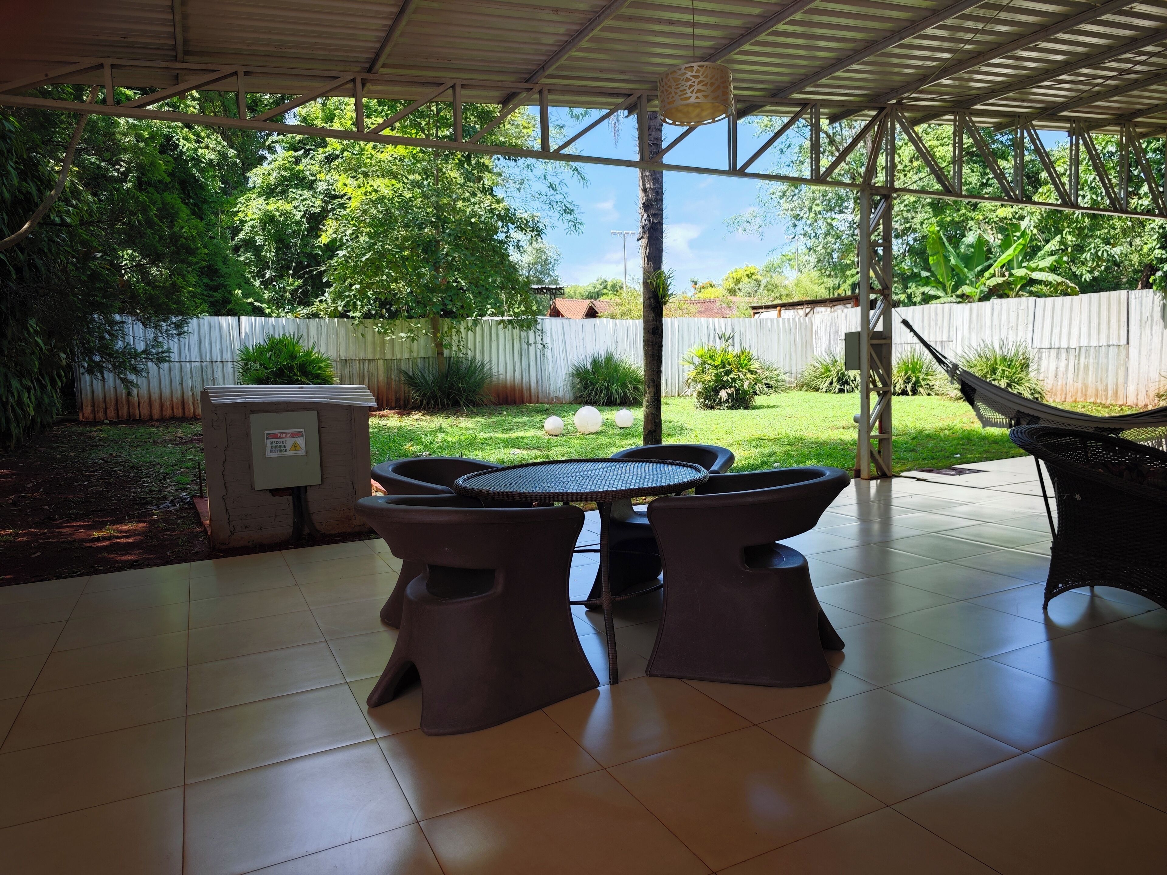 A covered patio with a round table and chairs, a hammock, and a view of a backyard with a wooden fence and greenery.