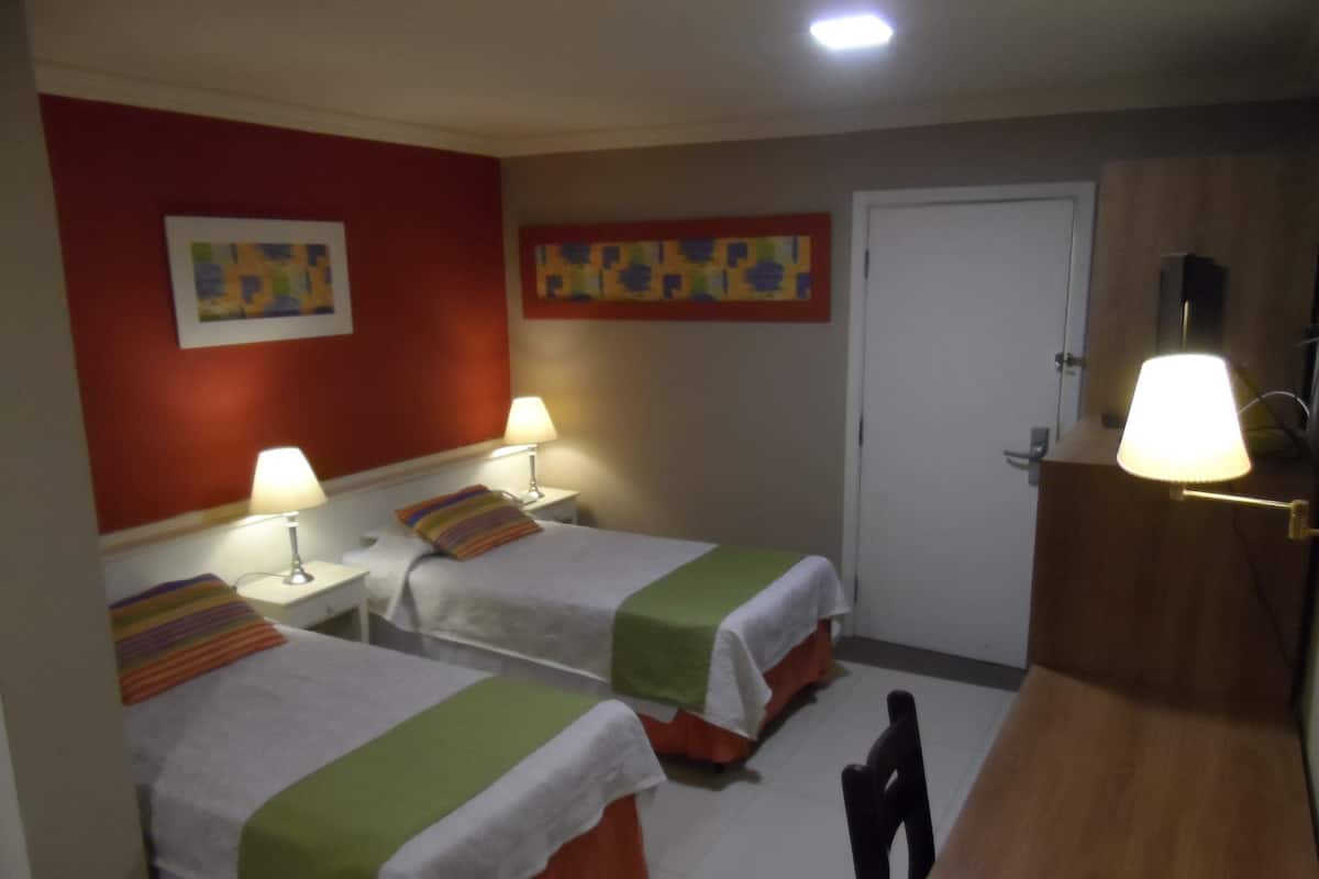 A hotel room with two beds, a desk, a chair, a door, and two wall-mounted lamps.