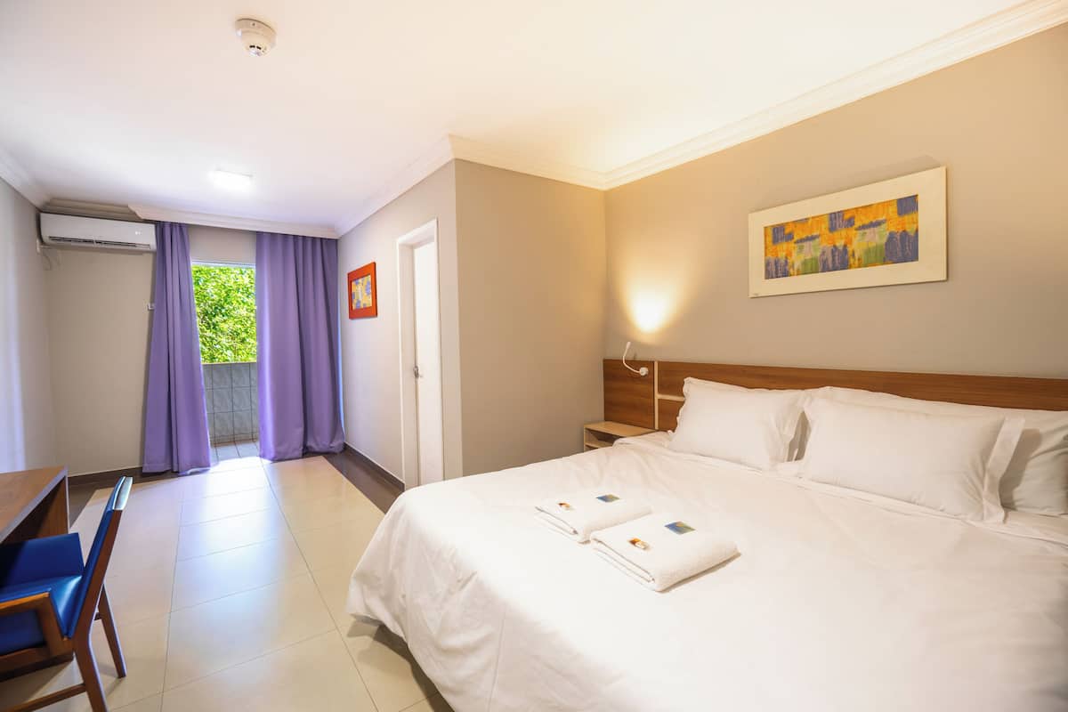 A hotel room with a bed, a desk, and a chair. There is a window with purple curtains, a painting on the wall, and an air conditioning unit on the wall.