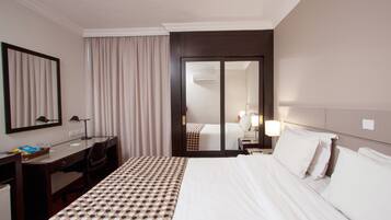 A hotel room with a bed, a desk, a chair, a mirror, a wardrobe, and a bathroom visible through a door.