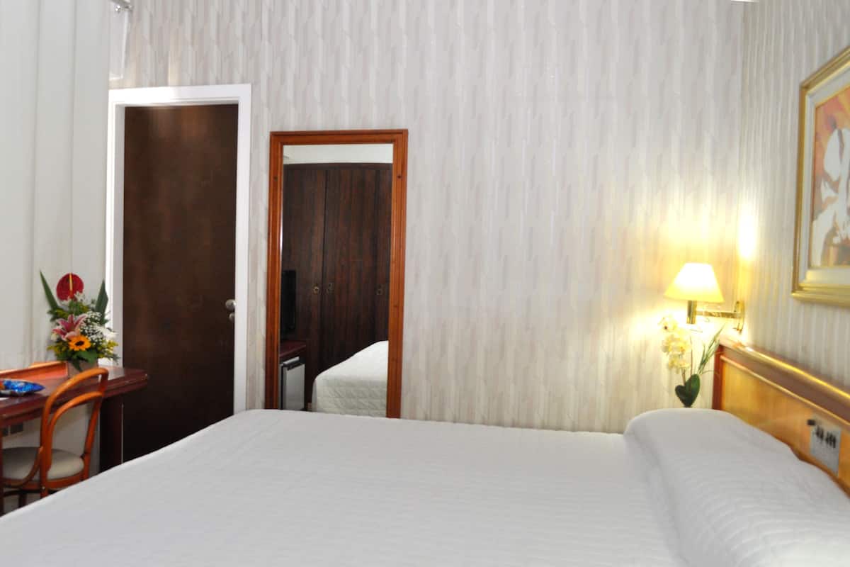 A hotel room with a bed, a desk with a chair, a lamp, and a door.