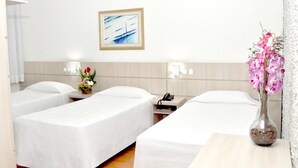 A hotel room with two single beds, a bedside table with a phone, a vase with flowers, and a painting on the wall. - Caravelle Palace Hotel (Curitiba)