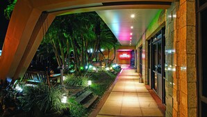 A well-lit pathway leading to a building with a neon sign, surrounded by greenery and palm trees. - Lira Hotel (Curitiba)
