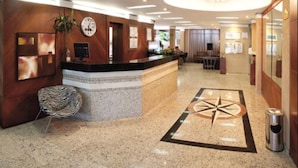 A hotel reception area with a reception desk, a clock, a plant, and a patterned floor design. - Lira Hotel (Curitiba)
