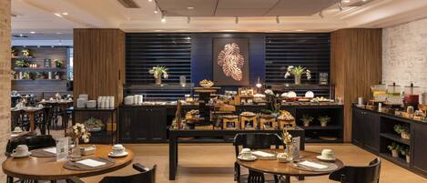 A modern cafe with wooden tables and chairs, a central counter with pastries, and a decorative wall with a leaf pattern.