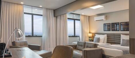 Habitación de hotel moderna con una cama grande, un escritorio y un área de descanso. Cuenta con ventanas amplias con cortinas, una lámpara de techo y un aire acondicionado.