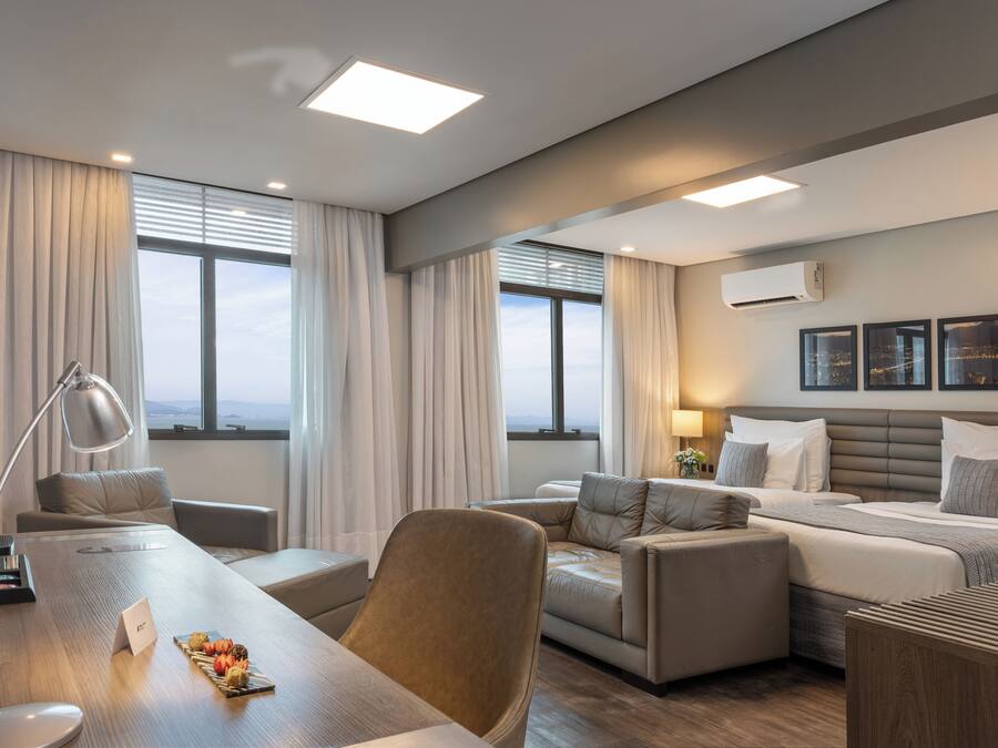 Une chambre d’hôtel moderne dotée d’un grand lit, d’un bureau et d’un coin salon. Elle dispose de grandes fenêtres avec des rideaux, d’un luminaire au plafond et d’un appareil de climatisation.