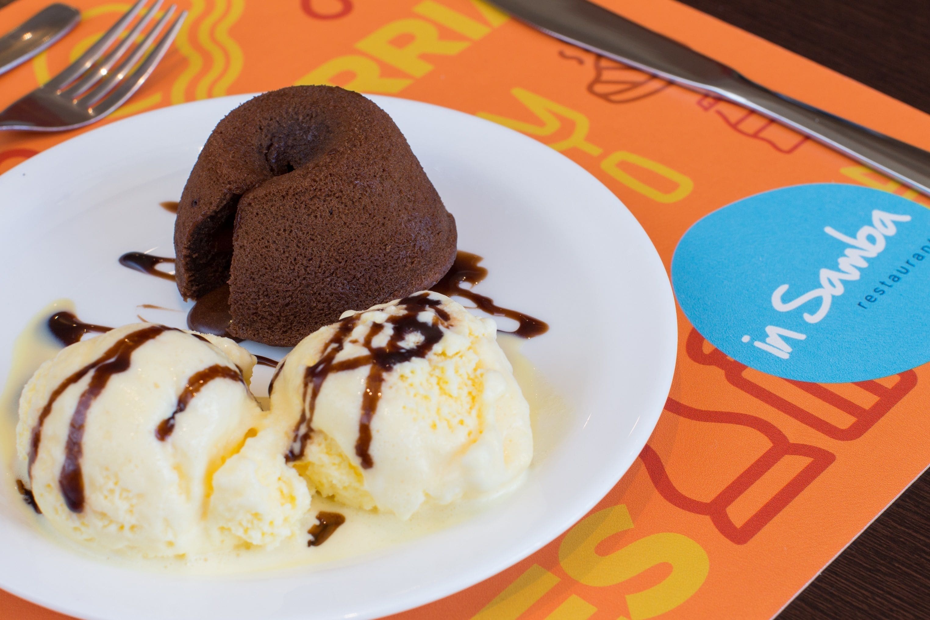 Un plato con pastel volcán de chocolate y helado de vainilla, servido sobre una fuente blanca con un logotipo azul.