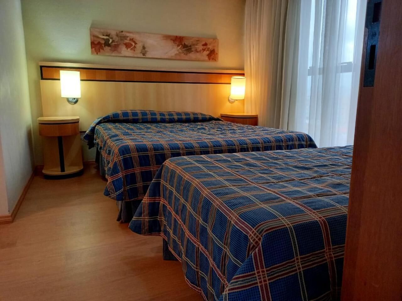 A hotel room with two beds, each with a plaid bedspread, a nightstand with a lamp, and a painting on the wall.