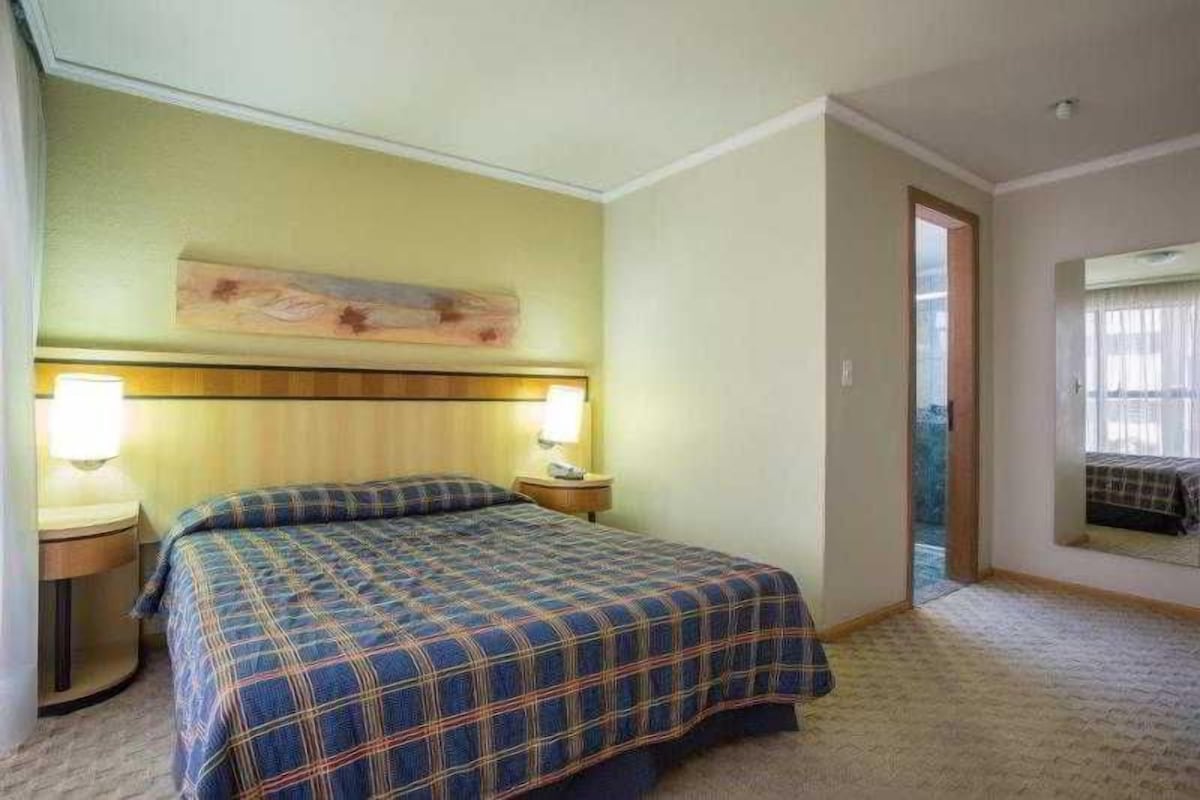Una habitació d'hotel amb un llit, dues làmpades de nit, un reposacapses i una finestra amb cortines.