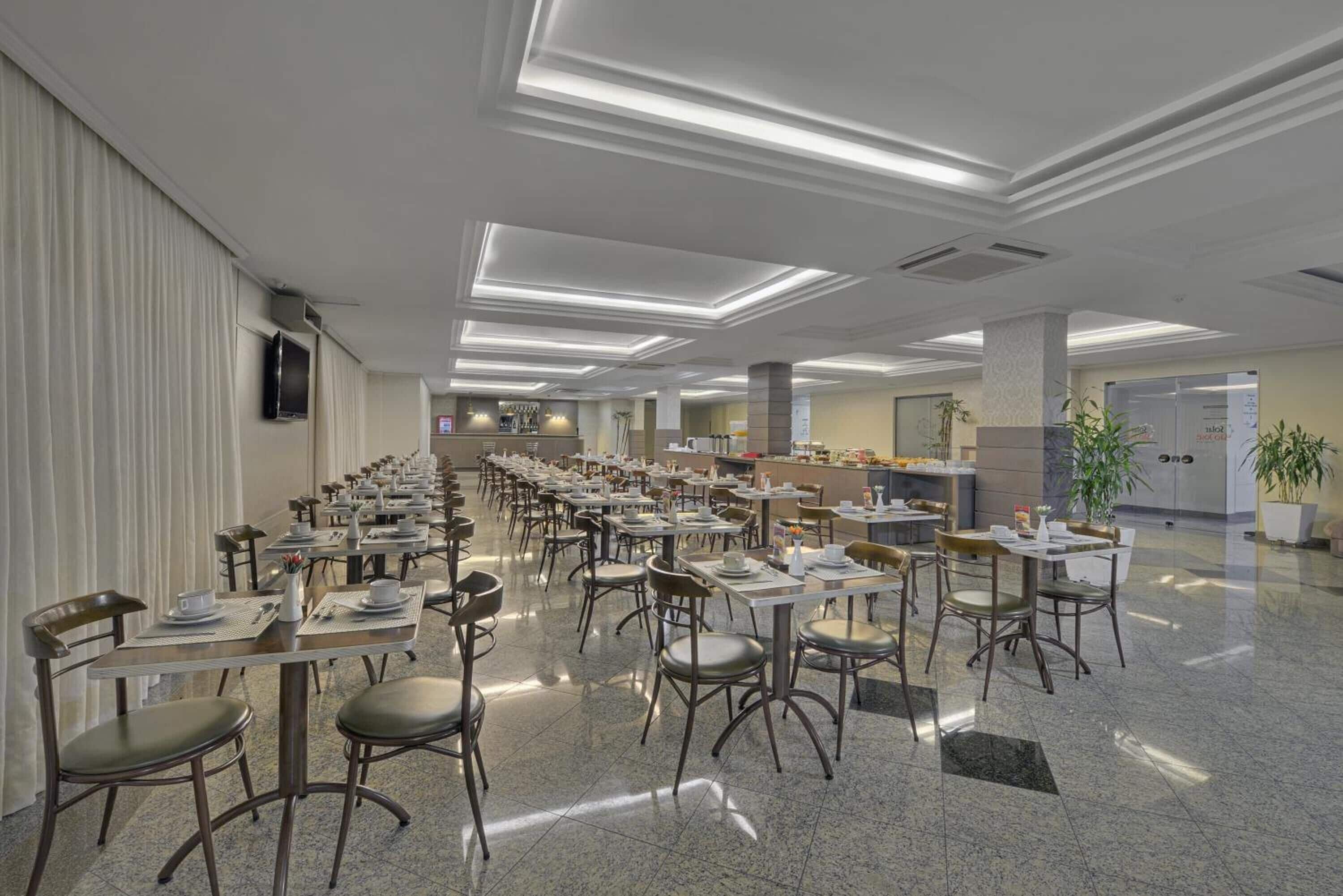 A spacious dining area with multiple tables and chairs, a kitchen in the background, and a modern ceiling design.