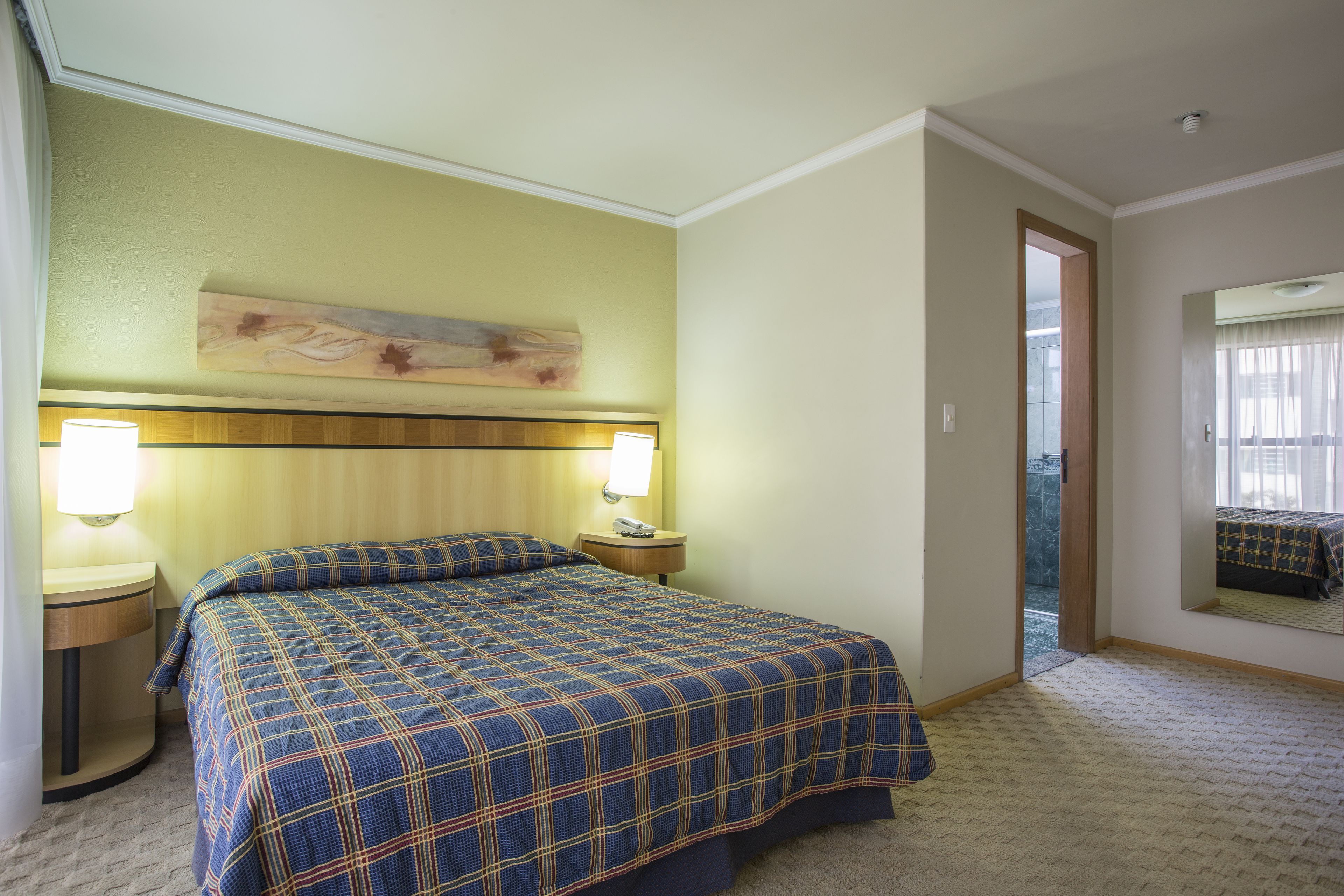 Habitació d'hotel amb un llit gran, dues làmpades de nit, un capçal de llit, un quadre sobre el llit, un mirall a la paret i una porta que dona a una altra habitació.