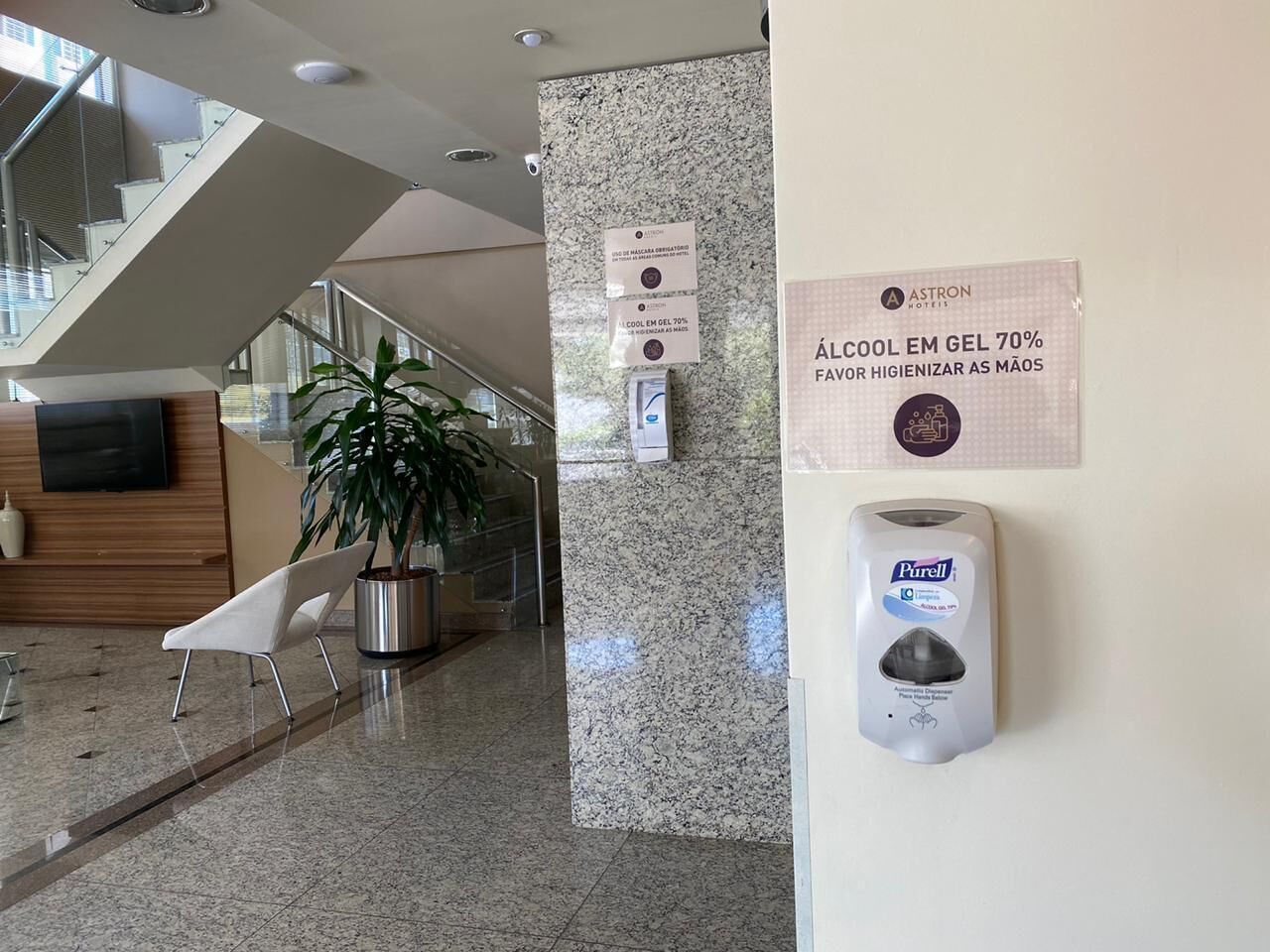 En hotellobby med modern design, inklusive en receptionsdisk, en sittgrupp med en stol och en krukväxt, en dispenser för handsprit och en skylt som marknadsför en handdesinfektionsgel med 70 % alkohol för god handhygien.