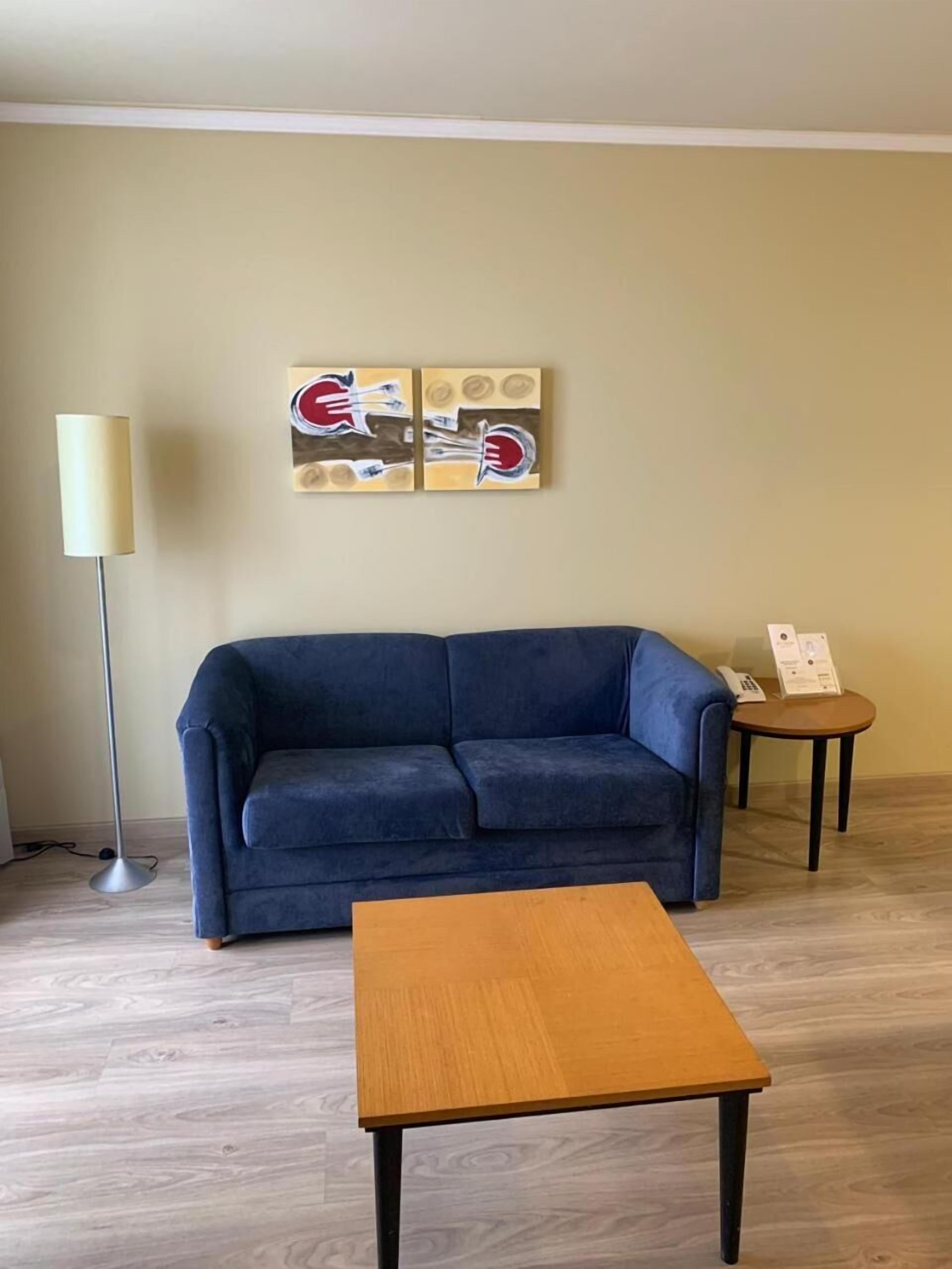 A living room with a blue sofa, a wooden coffee table, a floor lamp, and a small table with a phone and papers.
