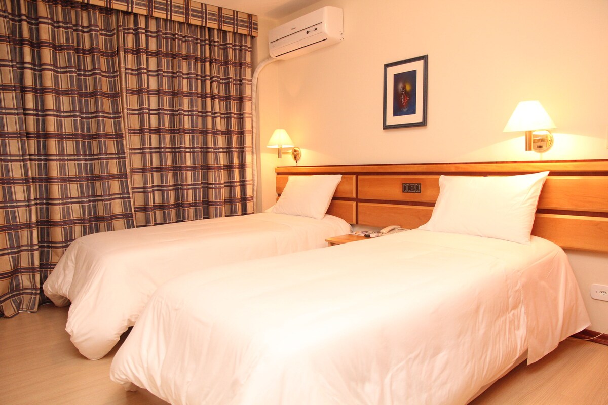 A hotel room with two beds, a wall-mounted air conditioner, two bedside lamps, a framed picture on the wall, and a plaid curtain.