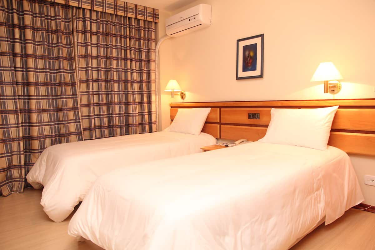A hotel room with two beds, a wall-mounted air conditioner, two bedside lamps, a framed picture on the wall, and a plaid curtain.