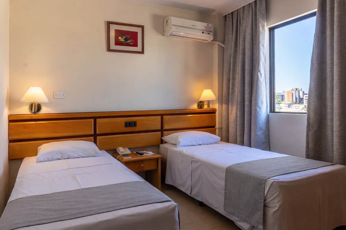 A hotel room with two single beds, a wooden headboard, bedside lamps, an air conditioning unit, a small table with a phone and remote, and a window with curtains.