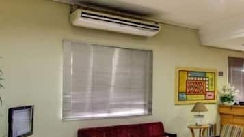 Sala de estar com sofá vermelho, poltronas listradas, mesa de centro de madeira e TV em um suporte. Um ar condicionado está fixado na parede, e há uma janela com persianas.