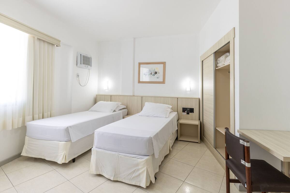 Une chambre d’hôtel avec deux lits simples, un bureau, une chaise, une armoire, une table de chevet, un climatiseur fixé au mur et un tableau encadré accroché au mur.