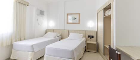 A hotel room with two single beds, a desk, a chair, a wardrobe, a bedside table, a wall-mounted air conditioner, and a framed picture on the wall.