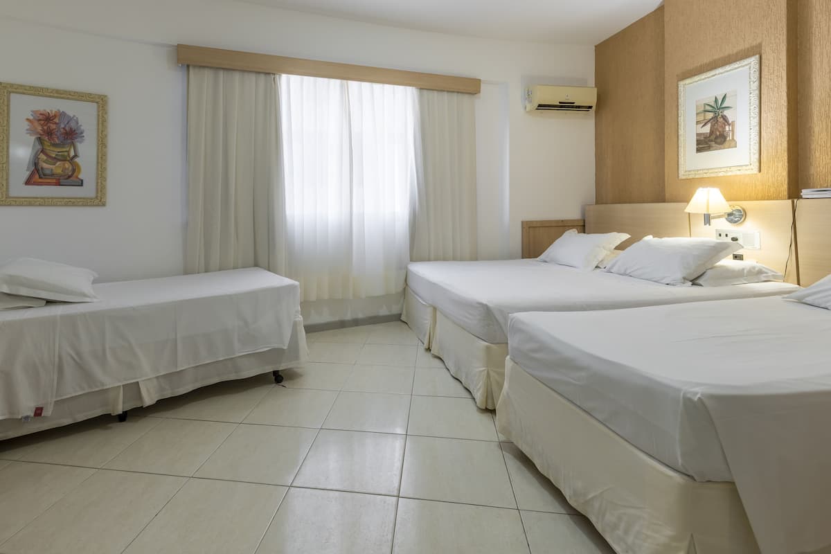 Une chambre d’hôtel avec deux lits simples, une grande fenêtre avec des rideaux, un climatiseur fixé au mur et des œuvres d’art encadrées.