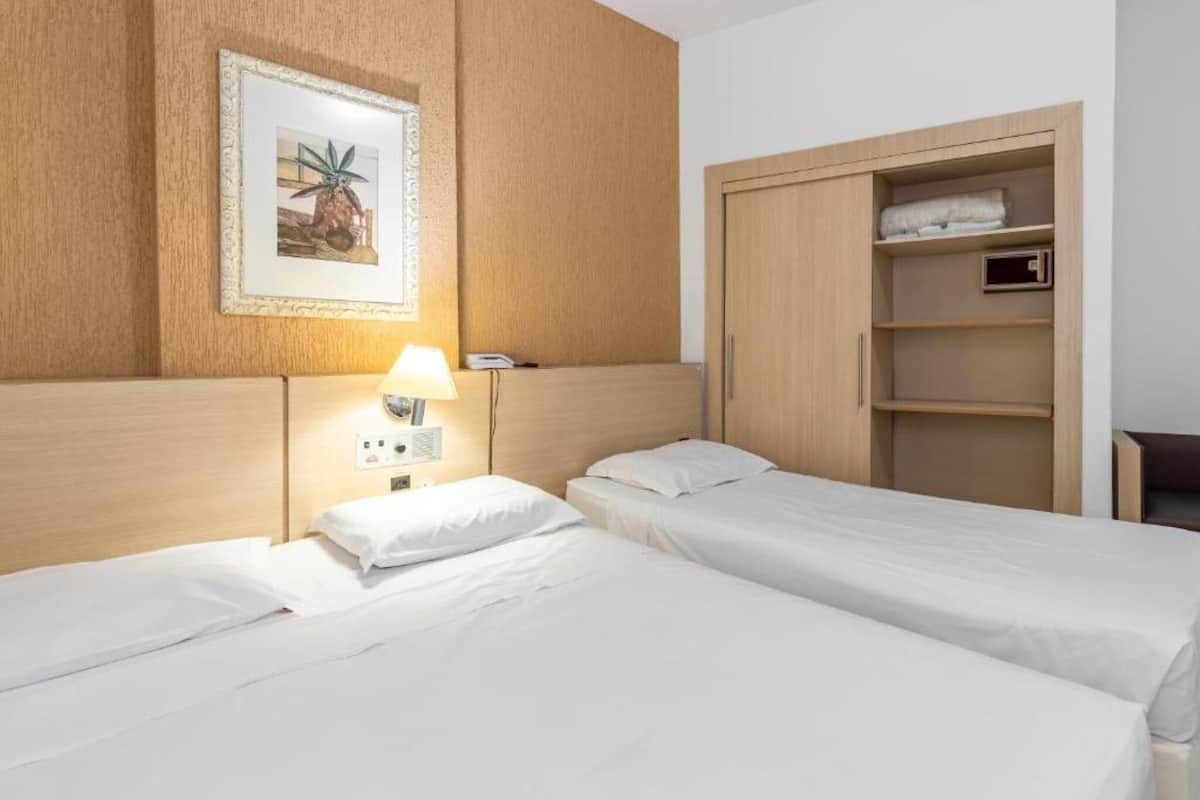 Une chambre d’hôtel avec deux lits, une tête de lit en bois, une lampe fixée au mur et une armoire avec des étagères.