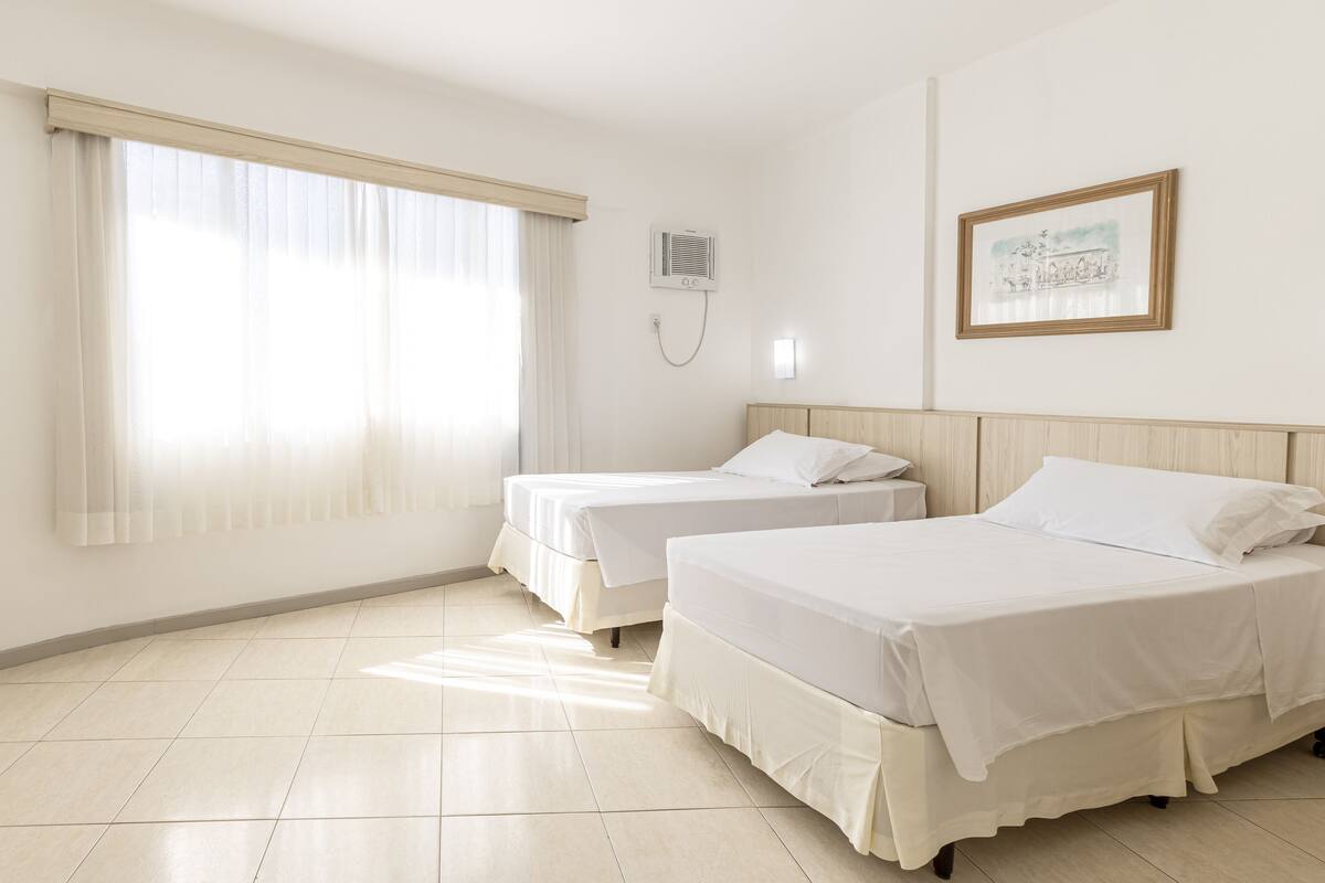 Une chambre d’hôtel avec deux lits, une grande fenêtre avec des rideaux légers, un climatiseur fixé au mur et un tableau encadré accroché au mur.