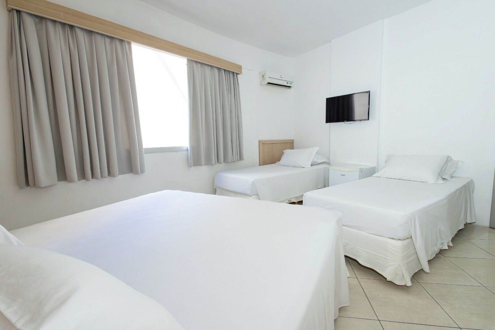 A hotel room with two beds, a TV, an air conditioning unit, and a window with curtains.