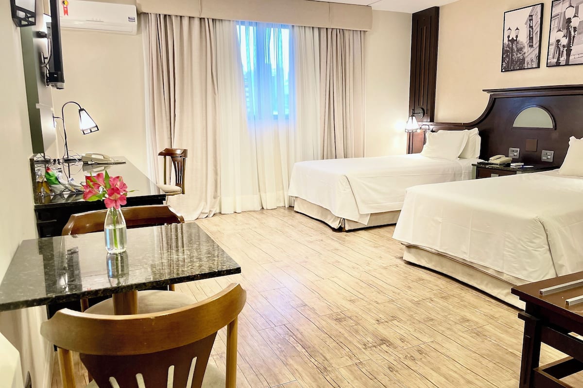 A hotel room with two beds, a desk with a chair, a small table with a vase, and a window with curtains.