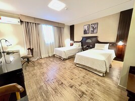 A hotel room with two beds, a desk with a lamp, a chair, and a large window with curtains.