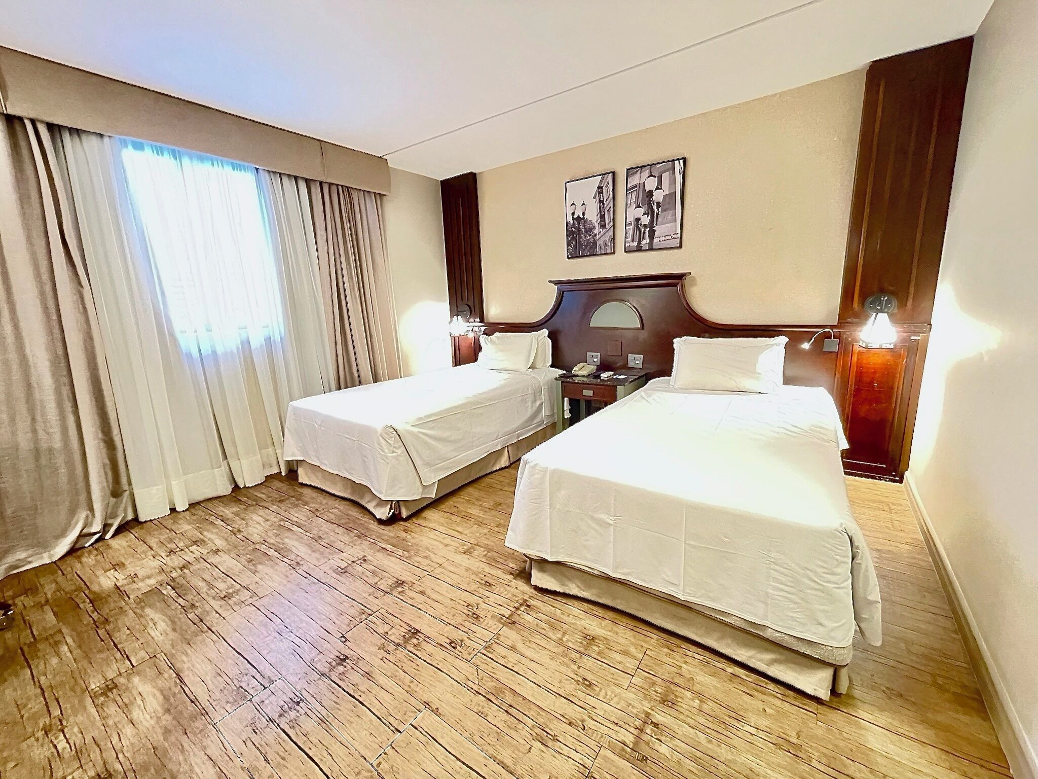 Habitación de hotel con dos camas individuales, suelo de madera, una mesita de noche con lámpara y una ventana con cortinas.