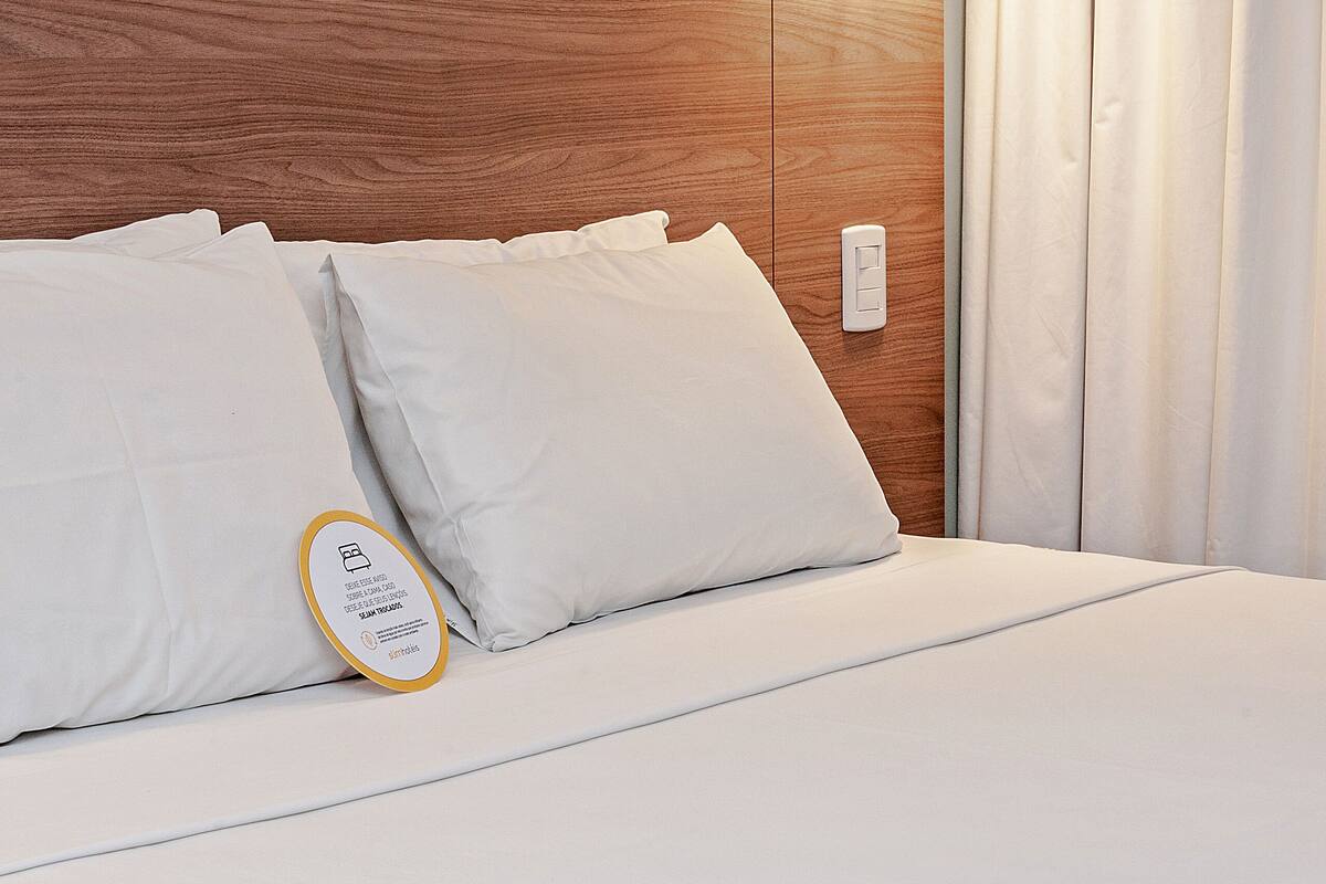 A neatly made bed with white linens, a wooden headboard, a wall-mounted lamp, and a power outlet.