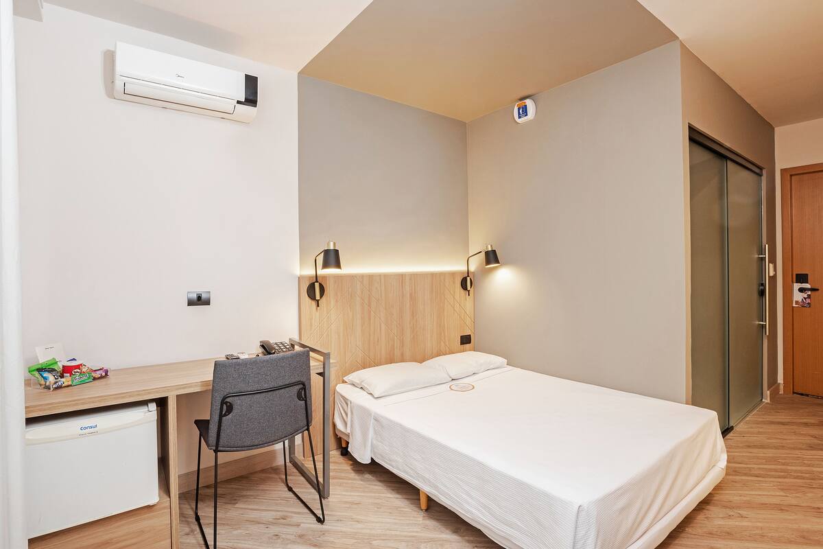 A hotel room with a bed, desk, chair, and an air conditioning unit.