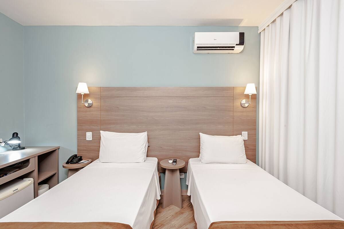 A hotel room with two beds, a wooden headboard, bedside lamps, a nightstand with a phone, and an air conditioning unit.