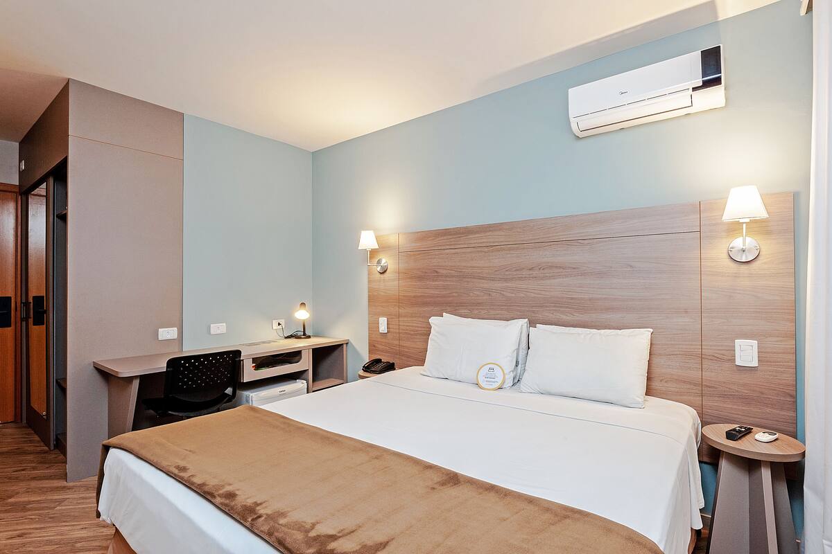 A hotel room with a bed, a desk, a chair, a nightstand, and an air conditioning unit.