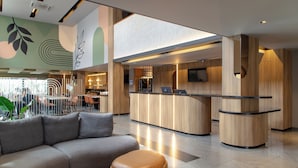 A modern reception area with a curved wooden counter, a seating area with a grey sofa and ottoman, and a wall with abstract leaf designs.
