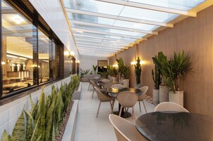 Un espacio exterior moderno para comer, con paredes de vidrio, mesas, sillas y plantas en macetas.
