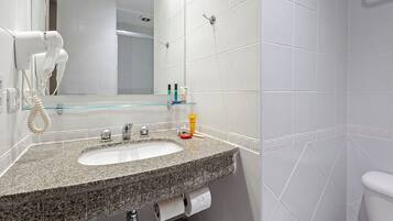 Un baño con lavamanos de granito, azulejos blancos, repisas de vidrio con artículos de aseo y un secador de pelo fijado en la pared.