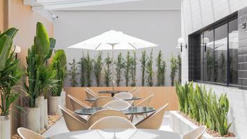 Um pátio moderno ao ar livre, com mesas redondas e cadeiras de vime, rodeado por plantas em vasos e um grande guarda-sol.