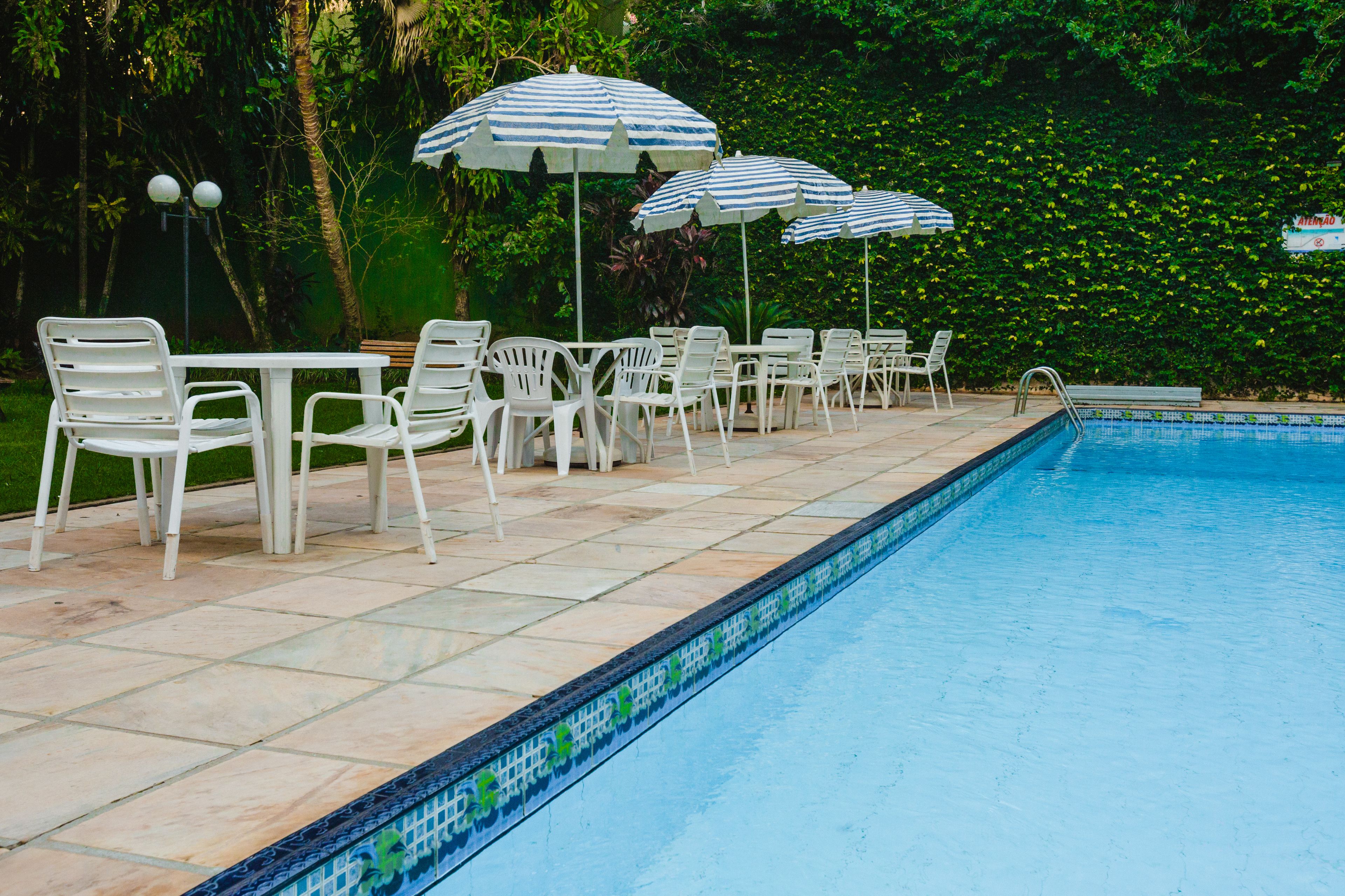 A poolside area with white chairs and striped umbrellas, surrounded by lush greenery.