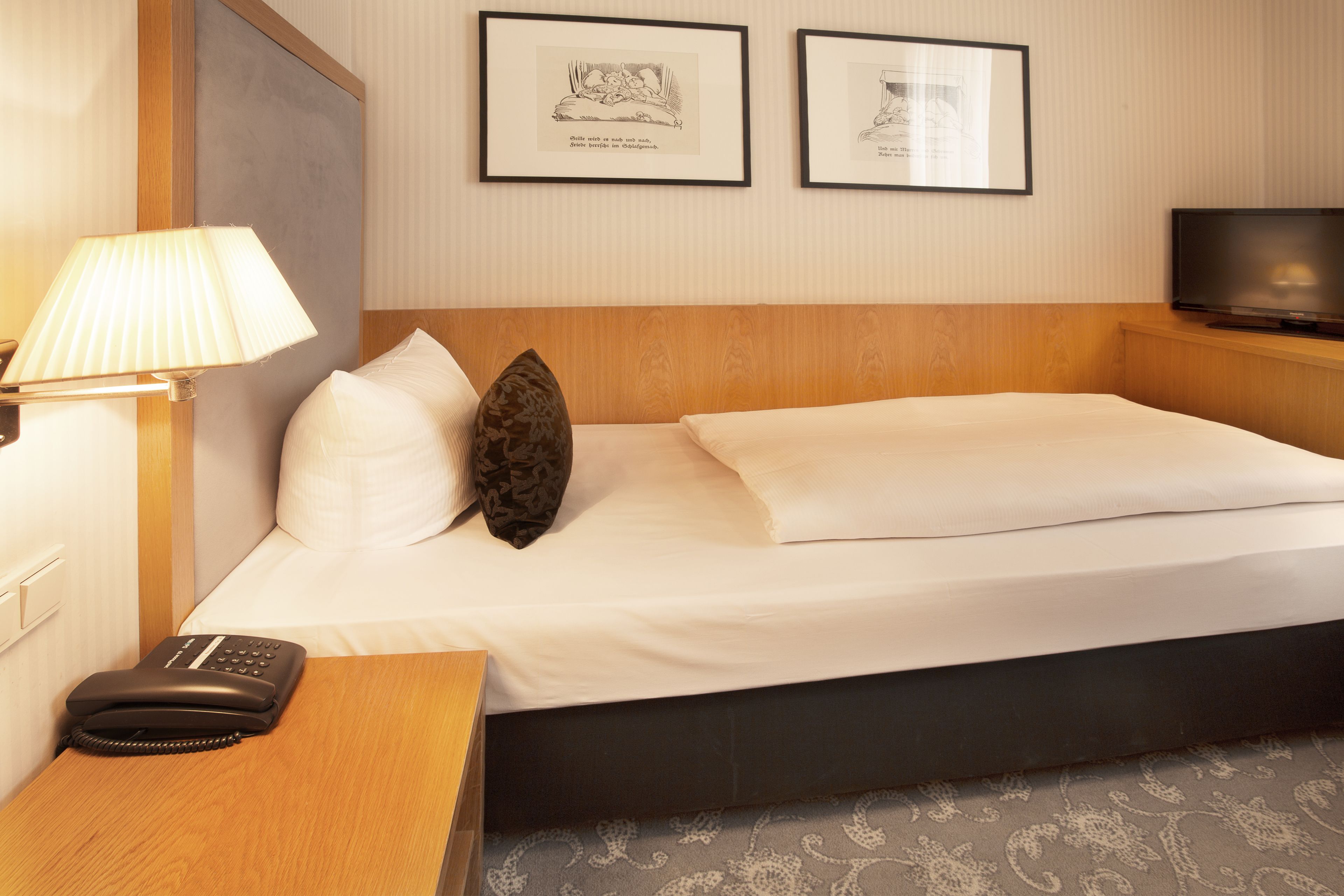 Una habitación de hotel con cama, lámpara de noche, teléfono y dos cuadros en la pared.